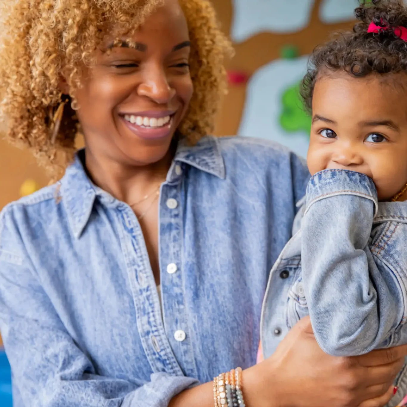 A smiling woman holding a baby girl who is playfully covering her mouth.